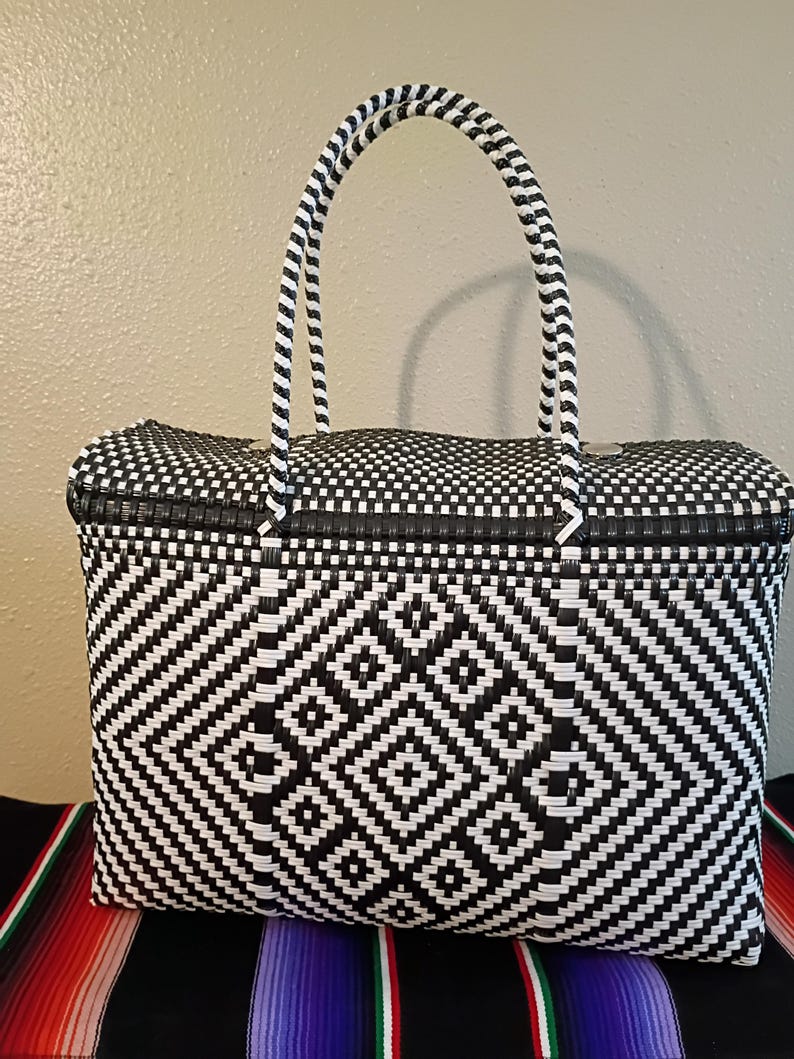 May include: Black and white woven tote bag with a geometric diamond pattern. The bag has two arched handles and a rectangular shape. The bag is sitting on a colorful striped textile.