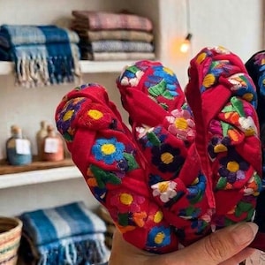 Puede incluir: Una mano sostiene varias diademas rojas con dise&ntilde;os florales bordados. Las diademas son el punto focal, con otros art&iacute;culos hechos a mano como cestas tejidas y textiles visibles en el fondo.