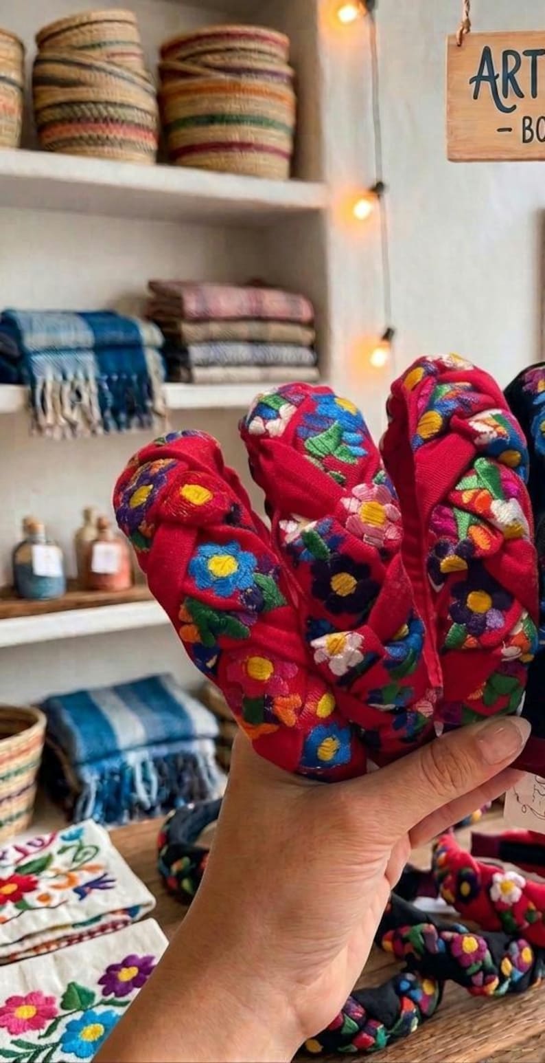 Puede incluir: Una mano sostiene varias diademas rojas con dise&ntilde;os florales bordados. Las diademas son el punto focal, con otros art&iacute;culos hechos a mano como cestas tejidas y textiles visibles en el fondo.