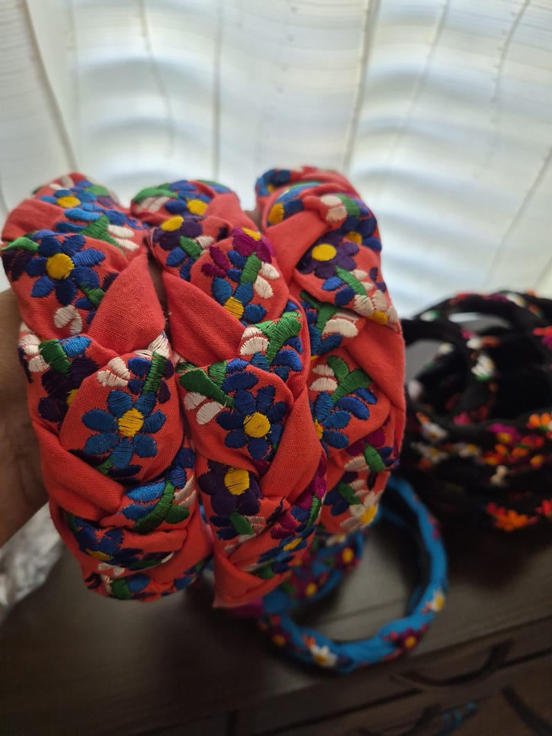 Puede incluir: Diademas trenzadas en tela coral, adornadas con patrones florales bordados en azul, amarillo y blanco. Tambi&eacute;n se ven diademas adicionales en azul y negro con dise&ntilde;os florales. Estos accesorios son perfectos para a&ntilde;adir un toque de color y estilo.