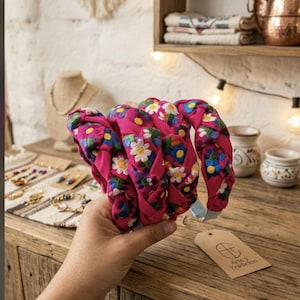 Puede incluir: Diademas trenzadas rosas con bordados florales blancos, azules y morados. Las diademas se sostienen en una mano, con una mesa de madera y objetos decorativos en el fondo. Una etiqueta con el texto "Arte y Tradici&oacute;n" es visible.