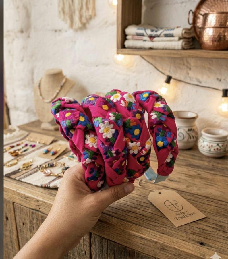 Puede incluir: Diademas trenzadas rosas con bordados florales blancos, azules y morados. Las diademas se sostienen en una mano, con una mesa de madera y objetos decorativos en el fondo. Una etiqueta con el texto "Arte y Tradici&oacute;n" es visible.