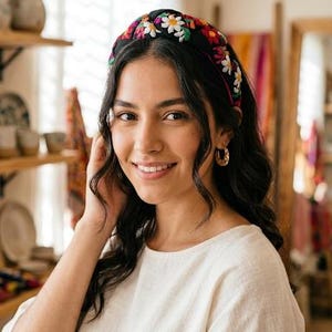 Puede incluir: Una mujer con una diadema negra bordada con flores coloridas, incluyendo margaritas blancas. La diadema es el punto focal, sobre el fondo del cabello oscuro de la mujer y una blusa color crema. Pendientes de aro dorados completan el look.