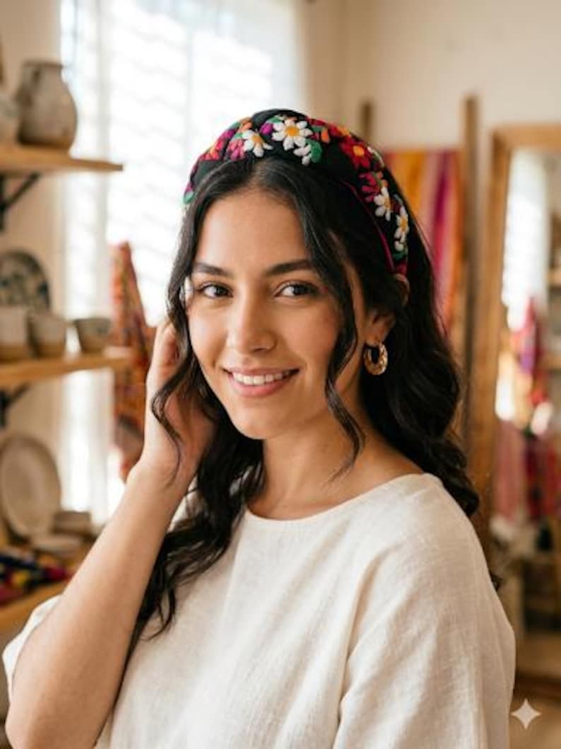 Puede incluir: Una mujer con una diadema negra bordada con flores coloridas, incluyendo margaritas blancas. La diadema es el punto focal, sobre el fondo del cabello oscuro de la mujer y una blusa color crema. Pendientes de aro dorados completan el look.