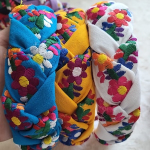 Puede incluir: Diademas trenzadas en azul, amarillo y blanco, cada una adornada con vibrantes bordados florales. Las coloridas flores presentan p&eacute;talos rojos, rosas, morados y blancos con hojas verdes. Estas diademas son un accesorio elegante.