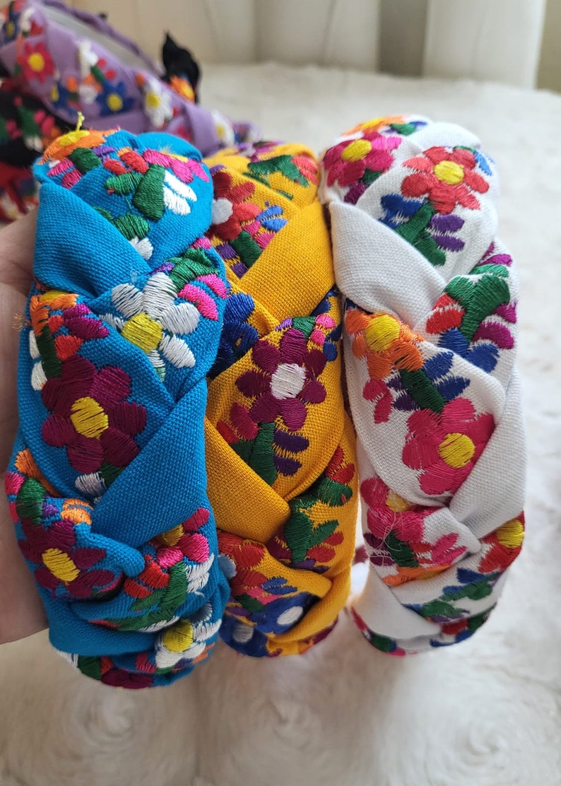 Puede incluir: Diademas trenzadas en azul, amarillo y blanco, cada una adornada con vibrantes bordados florales. Las coloridas flores presentan p&eacute;talos rojos, rosas, morados y blancos con hojas verdes. Estas diademas son un accesorio elegante.