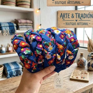 Puede incluir: Una diadema azul con un dise&ntilde;o trenzado, adornada con bordados florales coloridos. La diadema se sostiene frente a una tienda. El letrero de la tienda dice "ARTE & TRADICI&Oacute;N - BOUTIQUE ARTESANAL".