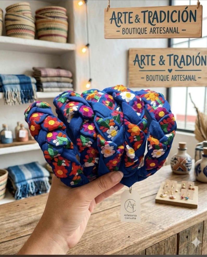 Puede incluir: Una diadema azul con un dise&ntilde;o trenzado, adornada con bordados florales coloridos. La diadema se sostiene frente a una tienda. El letrero de la tienda dice "ARTE & TRADICI&Oacute;N - BOUTIQUE ARTESANAL".