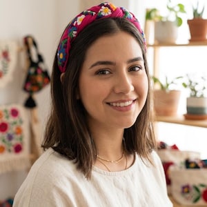 Puede incluir: Una diadema rosa brillante adornada con un colorido estampado floral. La diadema, anudada en la parte superior, es usada por una persona. El texto en la parte inferior dice: COLECCI&Oacute;N ARTESANAL - BORDADO A MANO.