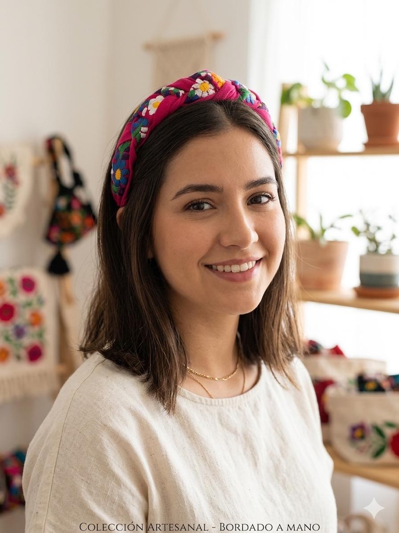 Puede incluir: Una diadema rosa brillante adornada con un colorido estampado floral. La diadema, anudada en la parte superior, es usada por una persona. El texto en la parte inferior dice: COLECCI&Oacute;N ARTESANAL - BORDADO A MANO.