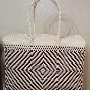 May include: A woven tote bag with a geometric diamond pattern in burgundy and cream. The bag has two braided handles and a rectangular shape. The top of the bag and the handles are cream-colored, while the body features a repeating diamond pattern.