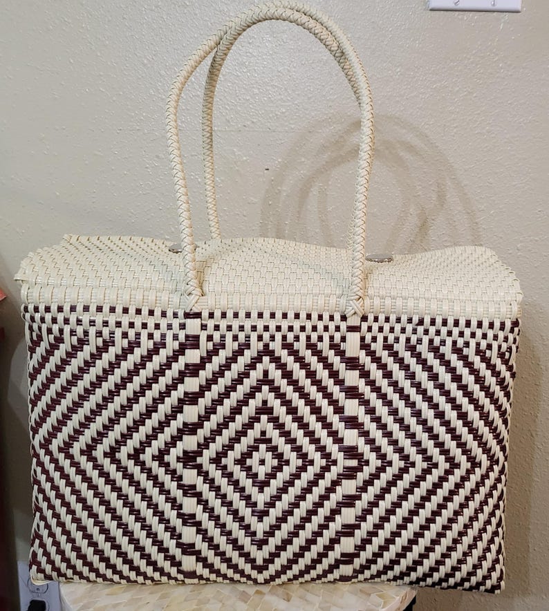 May include: A woven tote bag with a geometric diamond pattern in burgundy and cream. The bag has two braided handles and a rectangular shape. The top of the bag and the handles are cream-colored, while the body features a repeating diamond pattern.