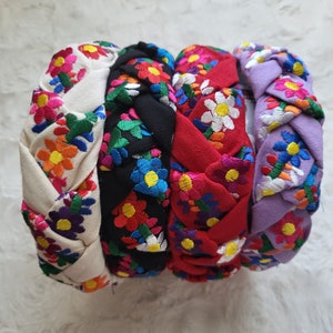 Puede incluir: Cuatro diademas trenzadas en crema, negro, rojo y lavanda, cada una adornada con bordados florales coloridos. Las flores presentan una mezcla de amarillo, naranja, rosa, azul y verde. Estas diademas son un accesorio elegante.