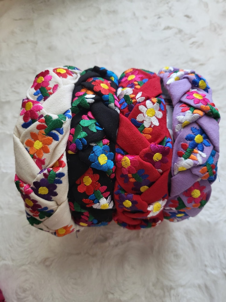 Puede incluir: Cuatro diademas trenzadas en crema, negro, rojo y lavanda, cada una adornada con bordados florales coloridos. Las flores presentan una mezcla de amarillo, naranja, rosa, azul y verde. Estas diademas son un accesorio elegante.
