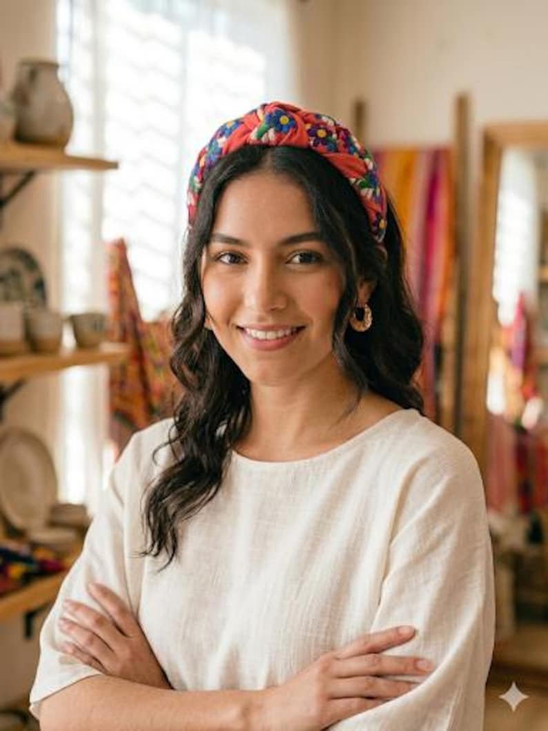 Puede incluir: Una mujer con una diadema roja con un estampado floral. La diadema tiene flores azules, amarillas y blancas. Lleva una blusa color crema y pendientes de aro dorados. Tiene los brazos cruzados.
