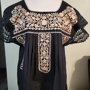 以下が含まれることがあります： スクエアネックと半袖が特徴の、黒色の刺繍チュニックトップ。胸と袖には、複雑なベージュの花柄刺繍が施されています。袖にはレースのトリムが付いています。チュニックはマネキンに展示されています。