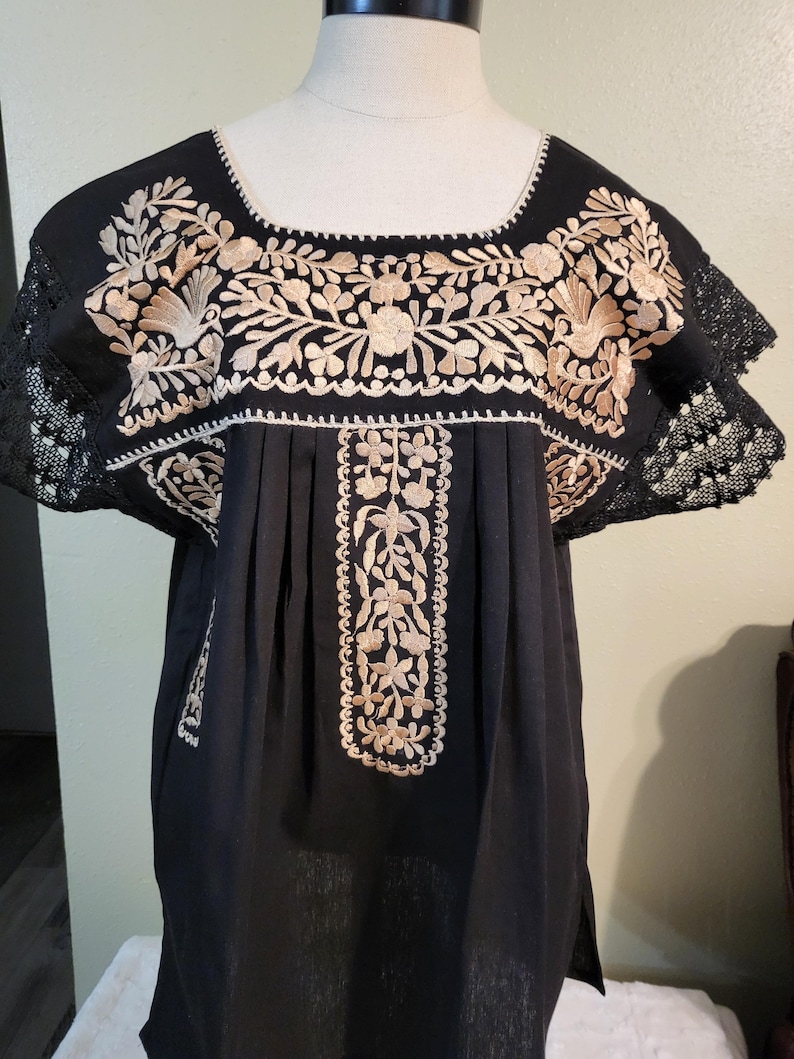 以下が含まれることがあります： スクエアネックと半袖が特徴の、黒色の刺繍チュニックトップ。胸と袖には、複雑なベージュの花柄刺繍が施されています。袖にはレースのトリムが付いています。チュニックはマネキンに展示されています。