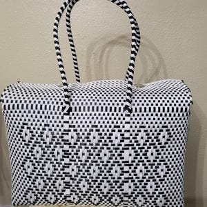 May include: A black and white woven tote bag with a rectangular shape and two arched handles. The bag features a repeating pattern of black and white squares and rectangles. The handles are black and white striped.