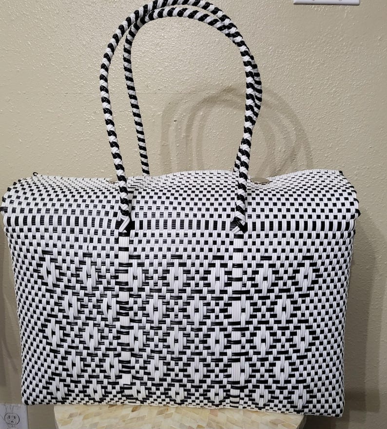 May include: A black and white woven tote bag with a rectangular shape and two arched handles. The bag features a repeating pattern of black and white squares and rectangles. The handles are black and white striped.
