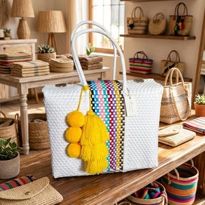 May include: A white woven tote bag featuring a vertical multicolored striped panel, two white braided handles, and a decorative yellow tassel accessory with three pom-poms. A small white rectangular tag is attached to one of the handles.