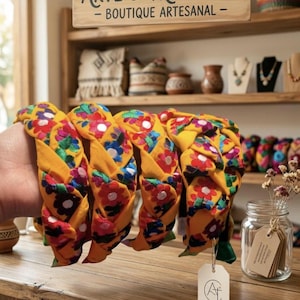 Puede incluir: Diademas trenzadas en tela amarilla vibrante, adornadas con bordados florales coloridos. Las diademas se exhiben sobre una superficie de madera, con un letrero que dice "ARTE & TRADICI&Oacute;N - BOUTIQUE ARTESANAL". Un frasco de flores secas y otros art&iacute;culos artesanales est&aacute;n en el fondo.