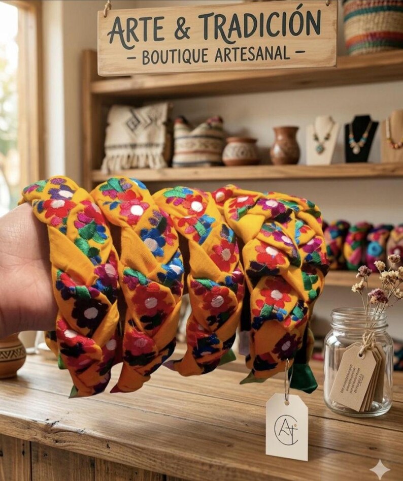 Puede incluir: Diademas trenzadas en tela amarilla vibrante, adornadas con bordados florales coloridos. Las diademas se exhiben sobre una superficie de madera, con un letrero que dice "ARTE & TRADICI&Oacute;N - BOUTIQUE ARTESANAL". Un frasco de flores secas y otros art&iacute;culos artesanales est&aacute;n en el fondo.