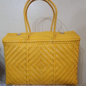 May include: A woven yellow tote bag with two braided handles. The bag features a diamond pattern on the front and a rectangular shape. The bag has a clasp closure and is made of a durable material.