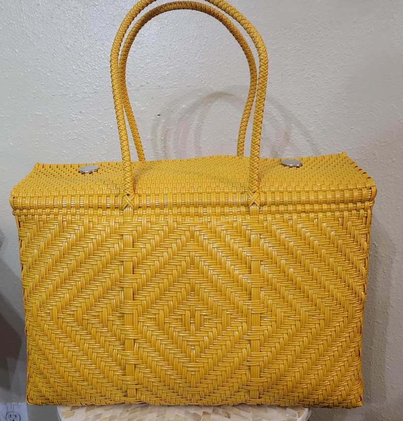 May include: A woven yellow tote bag with two braided handles. The bag features a diamond pattern on the front and a rectangular shape. The bag has a clasp closure and is made of a durable material.