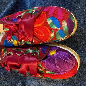 以下が含まれることがあります： 赤色の靴に、赤、紫、青、緑の色合いの花柄が刺繍されています。靴はバーガンディのリボンレースと織り込まれたソールが特徴です。