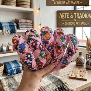 Puede incluir: Se muestran diademas trenzadas rosas con bordados florales multicolores. El letrero de la tienda dice "Arte & Tradicion - Boutique Artesanal."