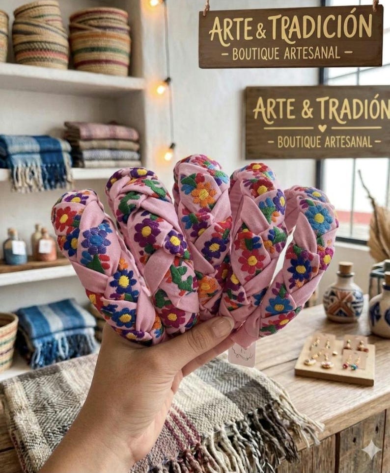 Puede incluir: Se muestran diademas trenzadas rosas con bordados florales multicolores. El letrero de la tienda dice "Arte & Tradicion - Boutique Artesanal."