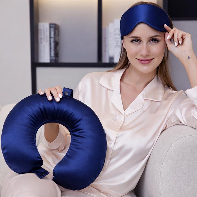 May include: A dark blue travel pillow and matching sleep mask. The pillow is U-shaped and made of a shiny, silky material. The sleep mask is also dark blue and covers the eyes. The items are designed for travel and relaxation.