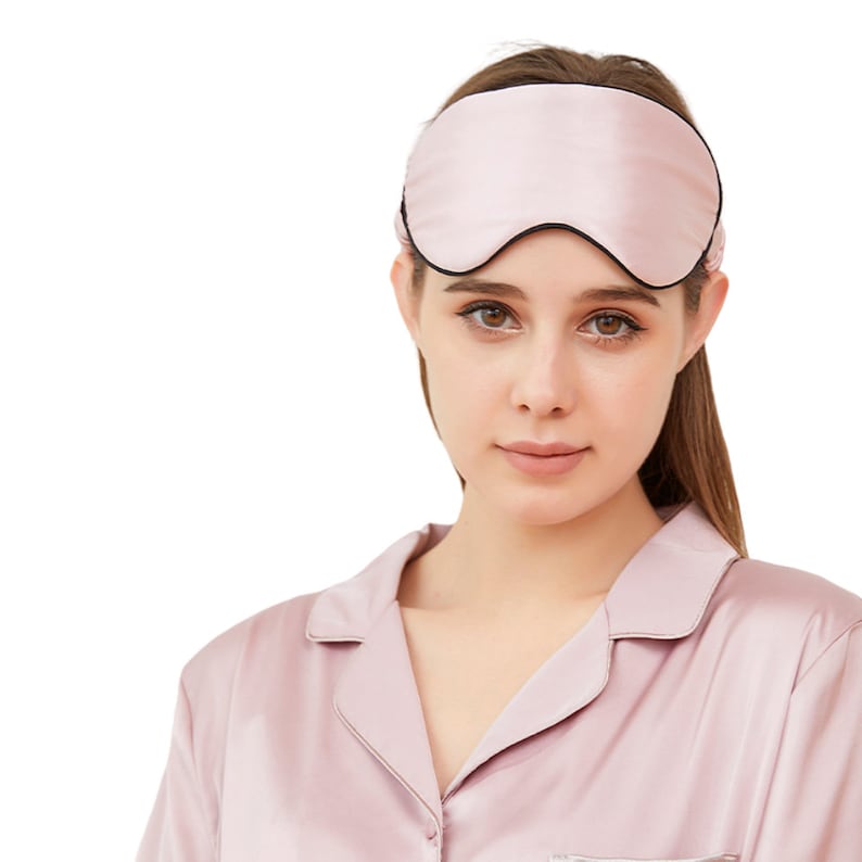 Op de afbeelding: Een lichtroze zijden slaapmasker met een zwarte rand. Het masker wordt gedragen door een persoon die een bijpassende lichtroze zijden pyjamajas draagt. Het slaapmasker is ontworpen om licht te blokkeren voor een betere slaap.