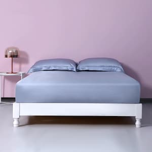 Puede incluir: Una sábana de cama de seda azul claro con dos fundas de almohada a juego sobre un marco de cama blanco con patas torneadas. La cama está frente a una pared rosa claro.