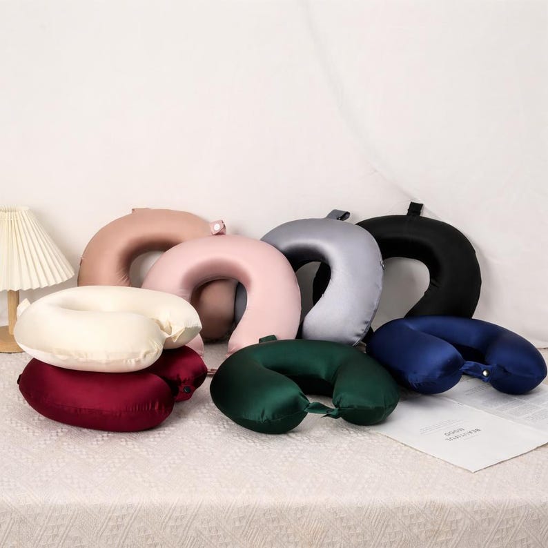 May include: A selection of U-shaped travel pillows in shades of cream, burgundy, pink, tan, grey, black, dark green, and navy blue. The pillows are made from a smooth, satin-like fabric, designed for comfort during travel and relaxation.