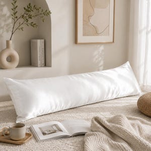 May include: A long, rectangular body pillow with a smooth, solid white fabric cover. The pillow is positioned horizontally across a textured surface, featuring a subtle sheen on the material. The design is simple, clean, and uniform in color.