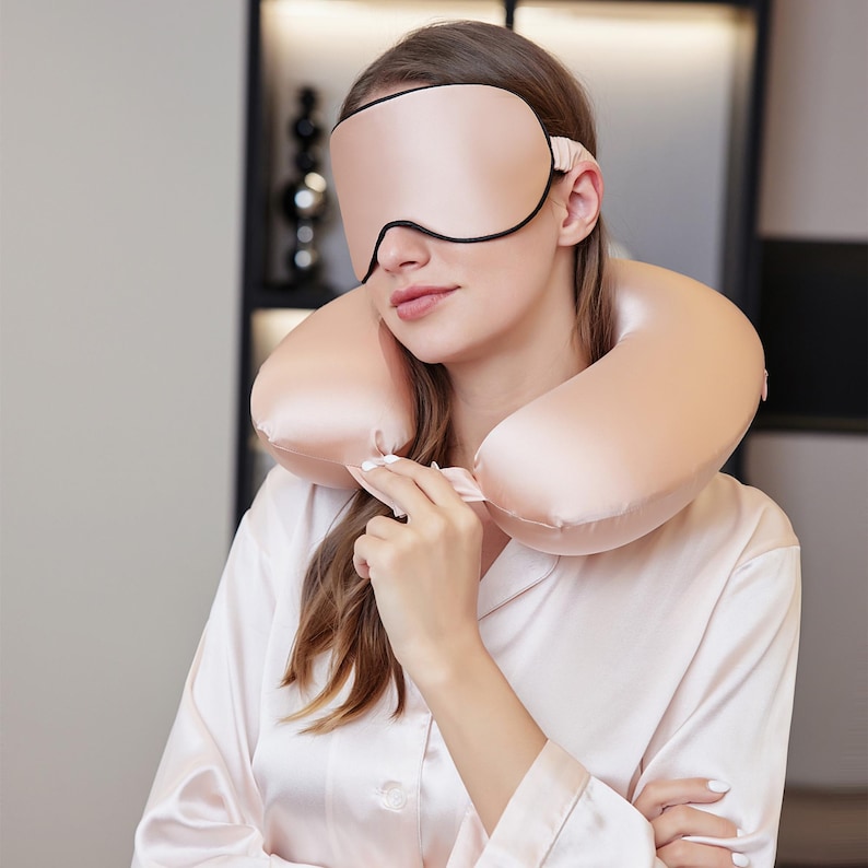 May include: A light pink sleep mask with black edging and a matching travel pillow. The travel pillow is around the neck. The woman is wearing a light pink satin pajama top.