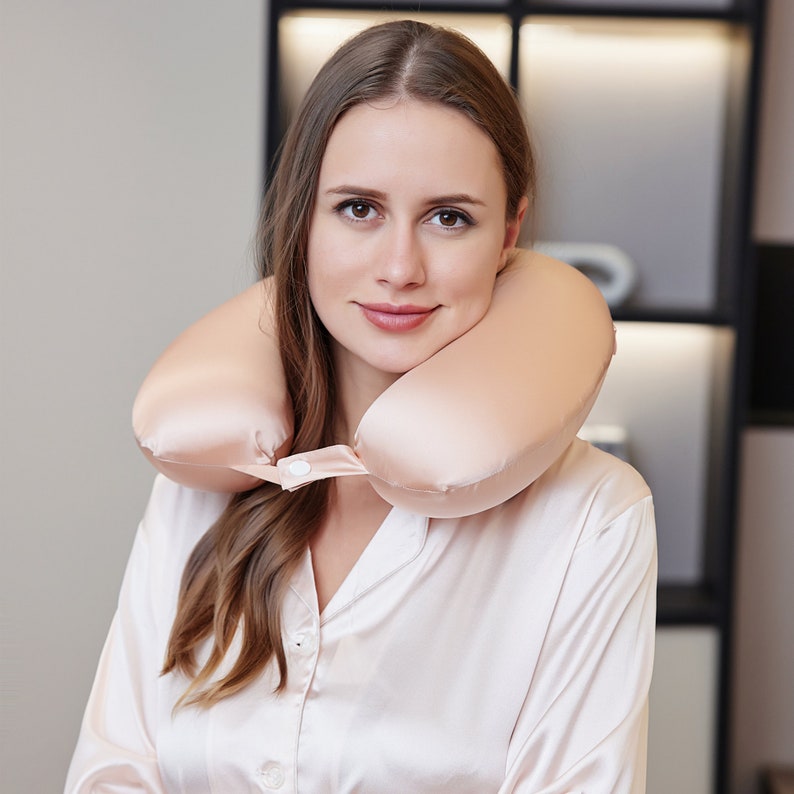 May include: A woman wearing a light pink satin shirt with a light pink travel neck pillow. The pillow is made of a soft, silky material and has a button closure.