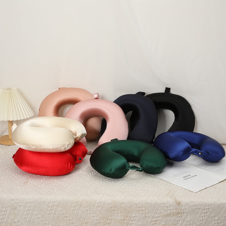 May include: Assortment of six travel neck pillows in various colors, including red, green, blue, cream, pink, and navy blue. The pillows are made of a soft, silky material and have a U-shape design.