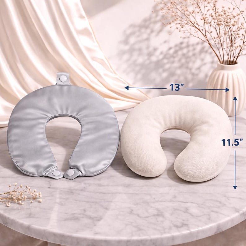 May include: Two travel neck pillows, one silver and one cream-coloured, are displayed on a marble surface. The silver pillow is 33 cm wide and 29 cm long. The cream-coloured pillow is 33 cm wide and 29 cm long. A vase with dried flowers is in the background.