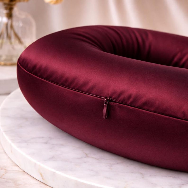 May include: A close-up of a burgundy-coloured, U-shaped pillow made of a smooth, satin-like material. A zip is visible along the seam. The pillow is resting on a white marble surface.