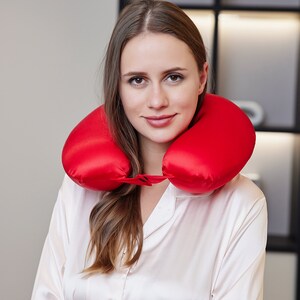 May include: A woman wearing a white shirt with a red travel neck pillow around her neck. The pillow is made of soft, silky material.