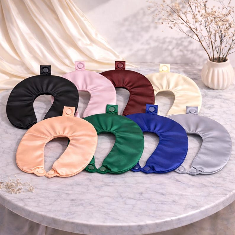 May include: A selection of satin travel neck pillows in a range of colours: black, pink, burgundy, cream, peach, green, blue, and grey. Each pillow has a button closure and is displayed on a marble surface.