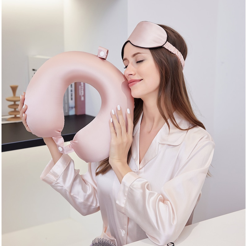 May include: A pink satin travel neck pillow with a strap and a button closure. The pillow is being held by a person wearing a pink satin sleep mask.