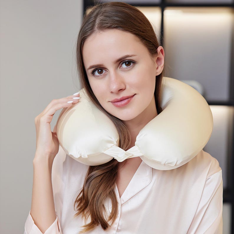 May include: A cream-coloured travel pillow, designed to support the neck. The pillow is made of a soft, satin-like material. The pillow is being worn around the neck.