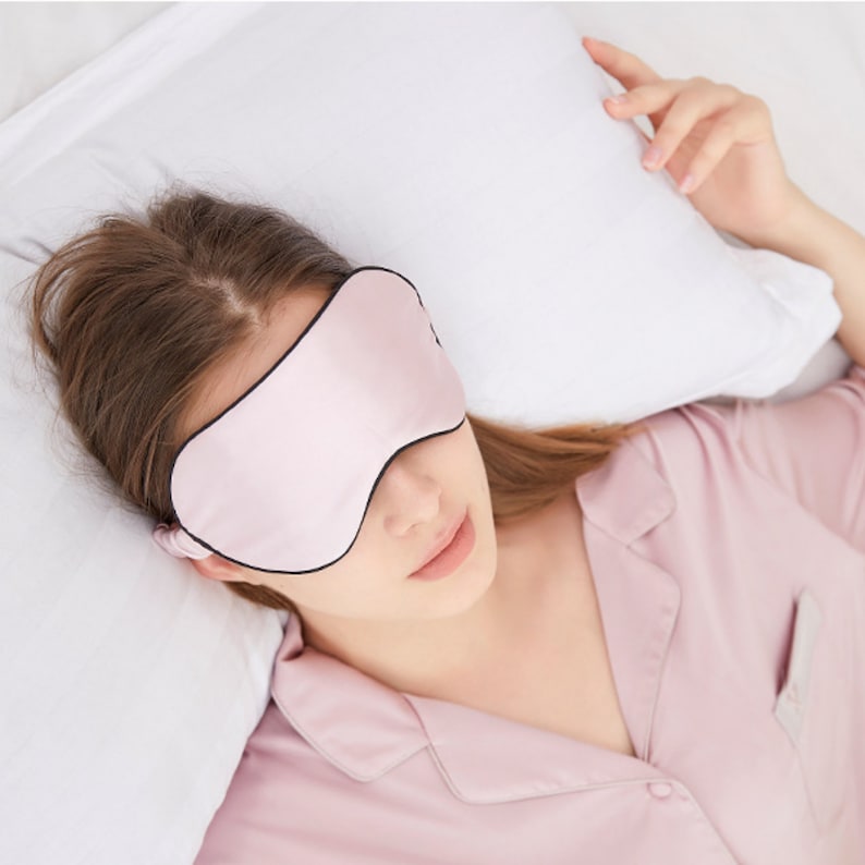 Op de afbeelding: Een lichtroze slaapmasker met een zwarte rand, gedragen door een persoon. De persoon draagt een lichtroze pyjamajas en ligt op een wit kussen. Het masker lijkt gemaakt van een zacht, zijdeachtig materiaal.