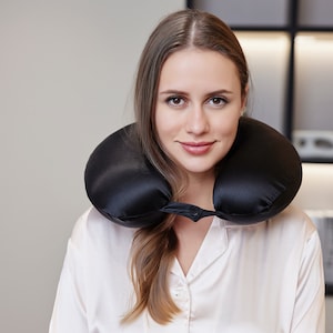 May include: A woman wearing a white shirt with a black travel pillow around her neck. She is looking directly at the camera and smiling.