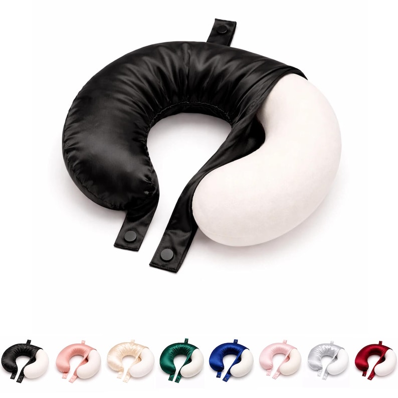 May include: A black and white travel pillow with a U-shape design. The black section is made of a shiny material, and the white section is a soft, plush fabric. The pillow has two black straps with snap closures. Several other colour variations are shown below.
