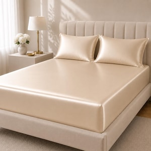 May include: A bed dressed with a beige satin sheet set and two matching pillowcases. The bed is made up and ready for use. The bed frame and headboard are also beige, creating a cohesive and elegant look. The room is well-lit, suggesting a bright and airy atmosphere.