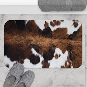 Puede incluir: Una alfombra de baño rectangular con estampado de piel de vaca marrón y blanca. La alfombra es suave y absorbente.